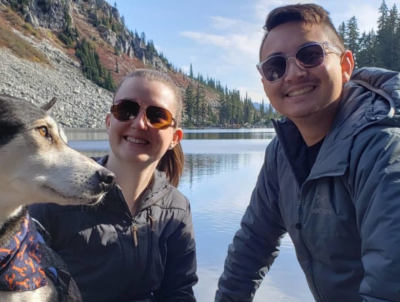 couple poses with their dog in front of lake and mountain landscape